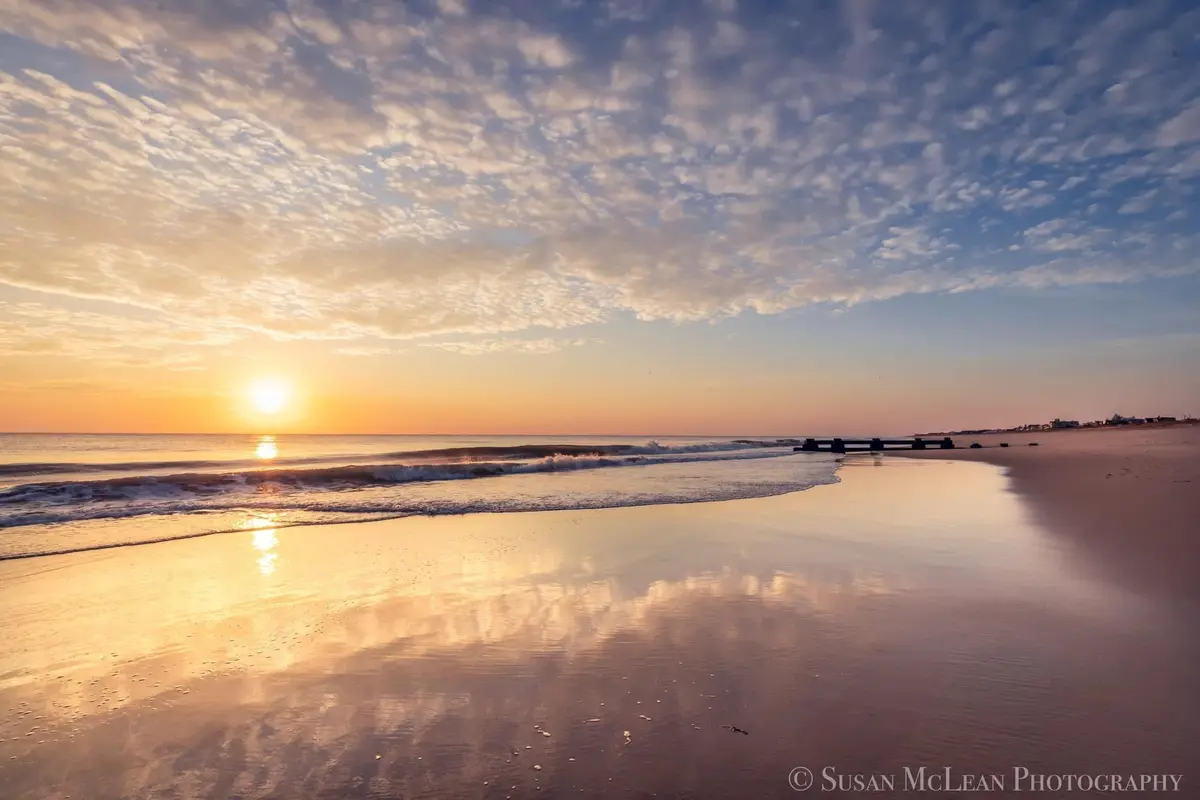 A serene beach scene at sunrise with the sun low on the horizon, casting golden light across the water and wet sand. The sky…