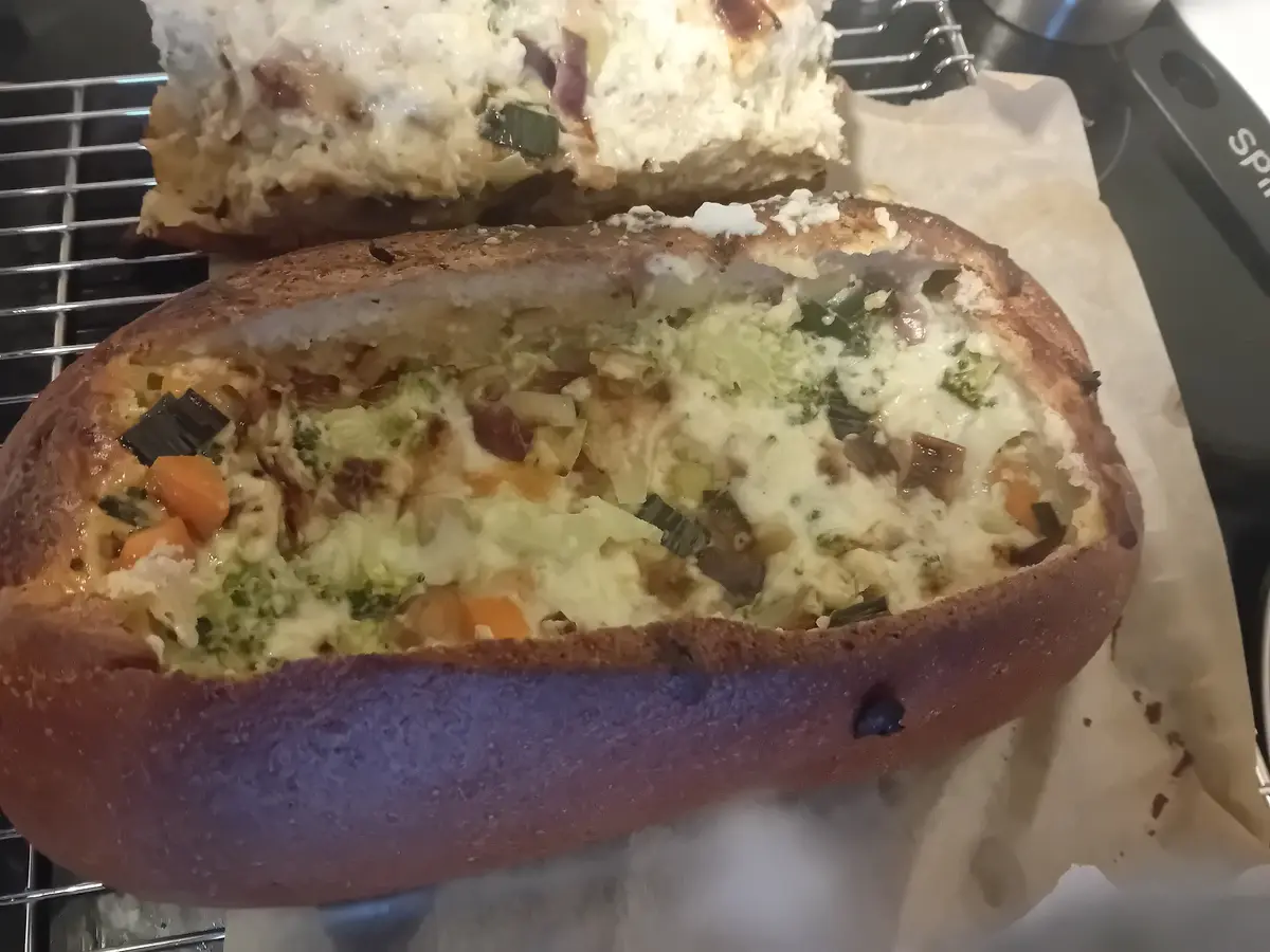 A baked potato, split open and filled with melted cheese, broccoli, carrots, and bacon bits, sits on parchment paper on a…