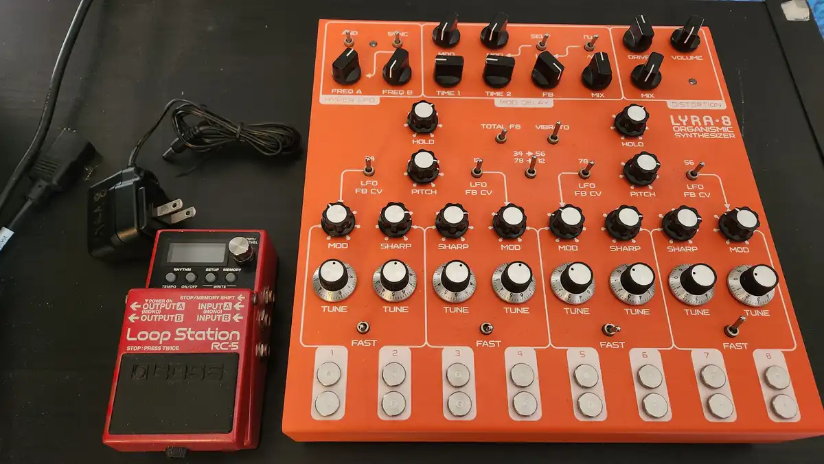 An orange Lyra-8 Organismic Synthesizer with numerous knobs and buttons sits next to a red Boss Loop Station RC-5 pedal and…