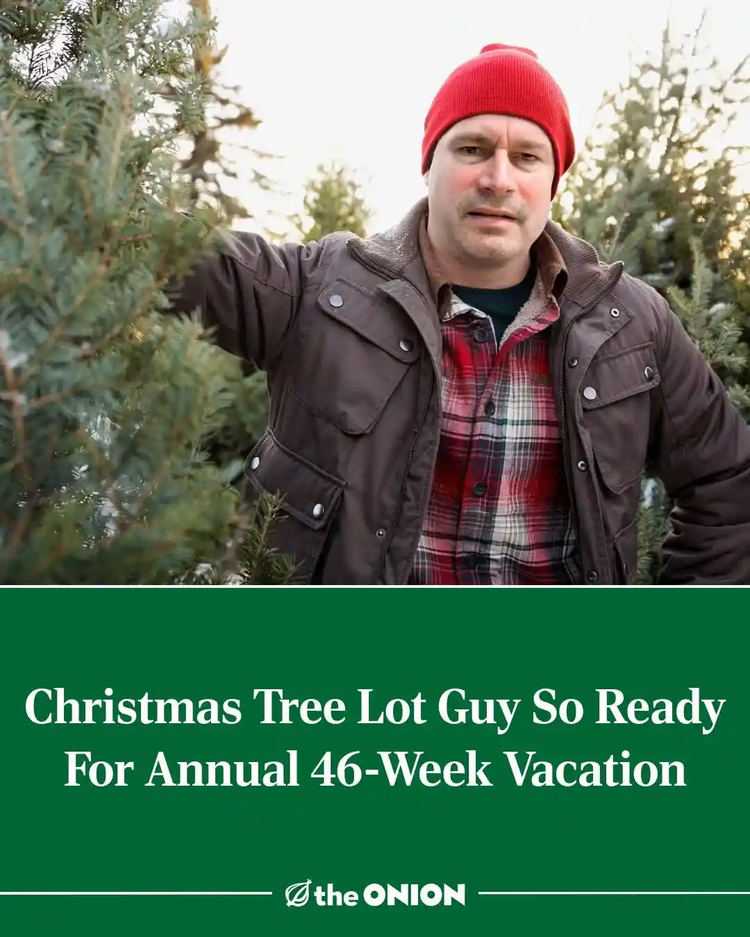 A man in a red beanie and brown jacket stands among evergreen trees, looking directly at the camera with a slightly weary…