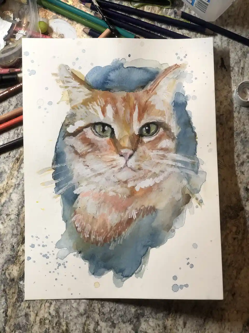 A watercolor painting of a fluffy orange and white cat's face on white paper, surrounded by blue watercolor splatters, sits…
