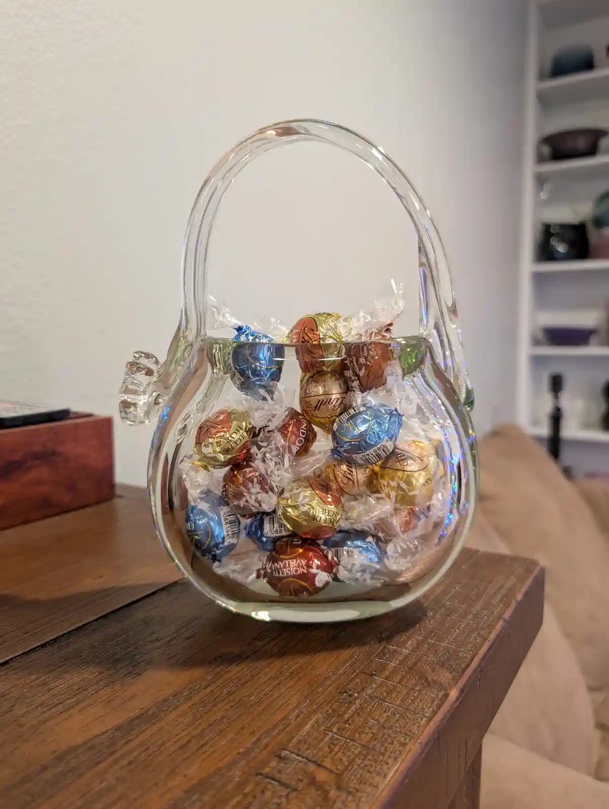 A clear glass candy dish shaped like a basket, filled with Lindt and other foil-wrapped chocolate eggs, sits on a wooden…