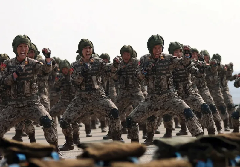 A group of soldiers in camouflage uniforms and helmets are performing a synchronized, energetic dance move with arms raised…
