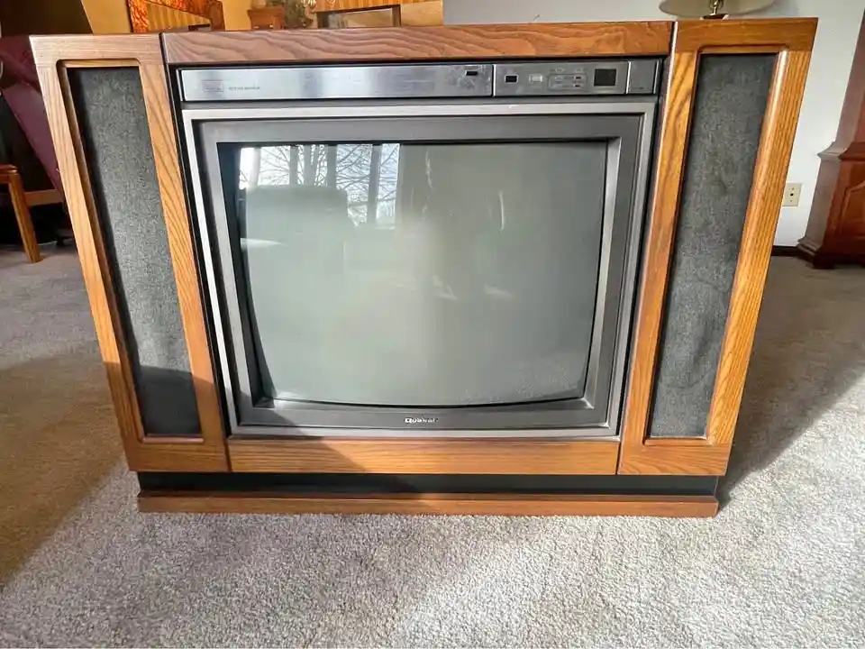A vintage wooden console television with a large, blank, reflective screen sits on a carpeted floor. The TV has a silver…