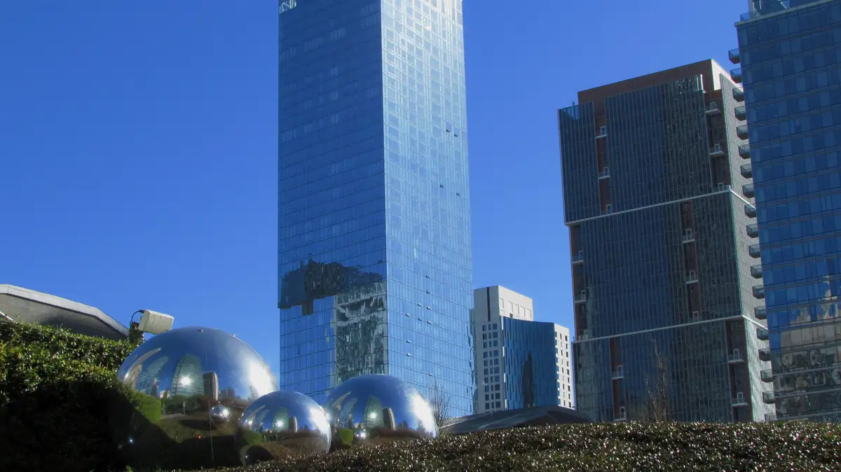 A photo of three large, shiny chrome spheres in a landscaped urban area, reflecting surrounding glass skyscrapers and a…