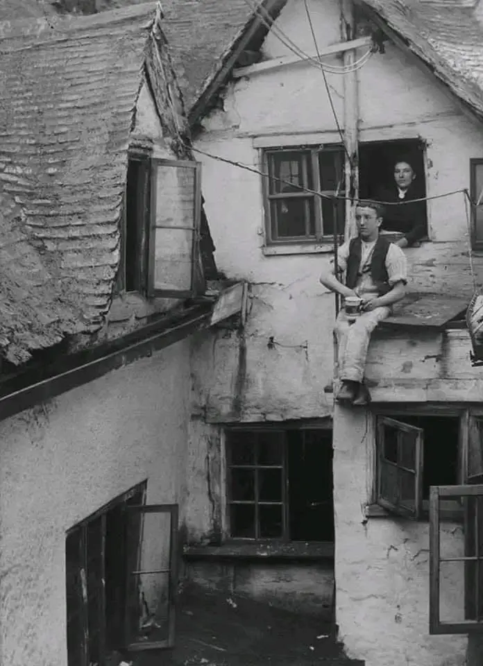 A black-and-white historical photo shows a man sitting precariously on a narrow wooden ledge between two old buildings…