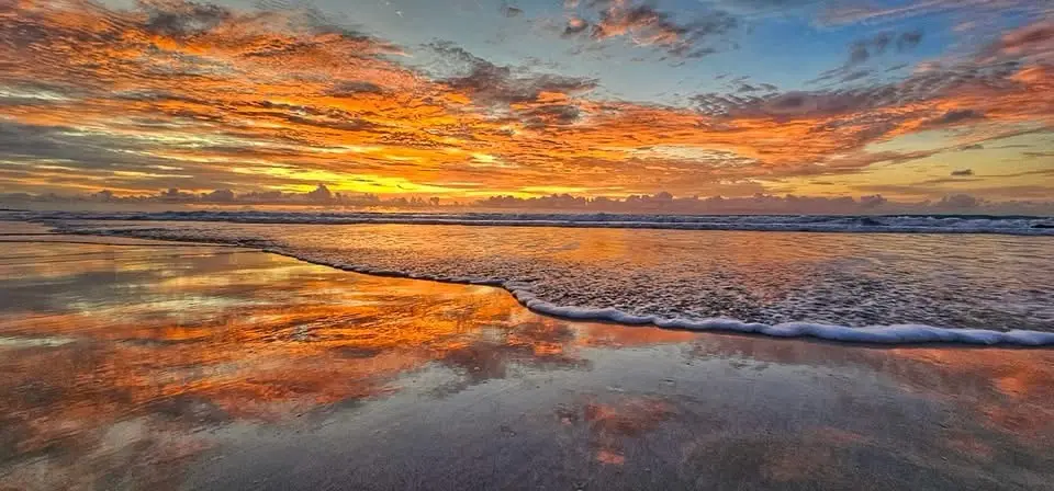 A vibrant, high-contrast photograph of a beach at sunset, with fiery orange and yellow clouds reflected in the wet sand as…