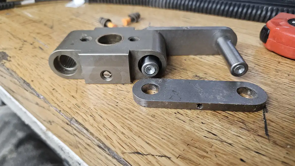 A close-up photo shows two custom-made metal mechanical parts resting on a worn wooden workbench, surrounded by workshop…