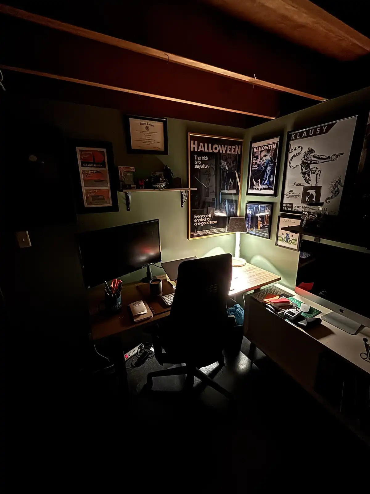 A dimly lit home office with a desk, chair, and computer, illuminated by a single lamp. The walls are covered in framed…
