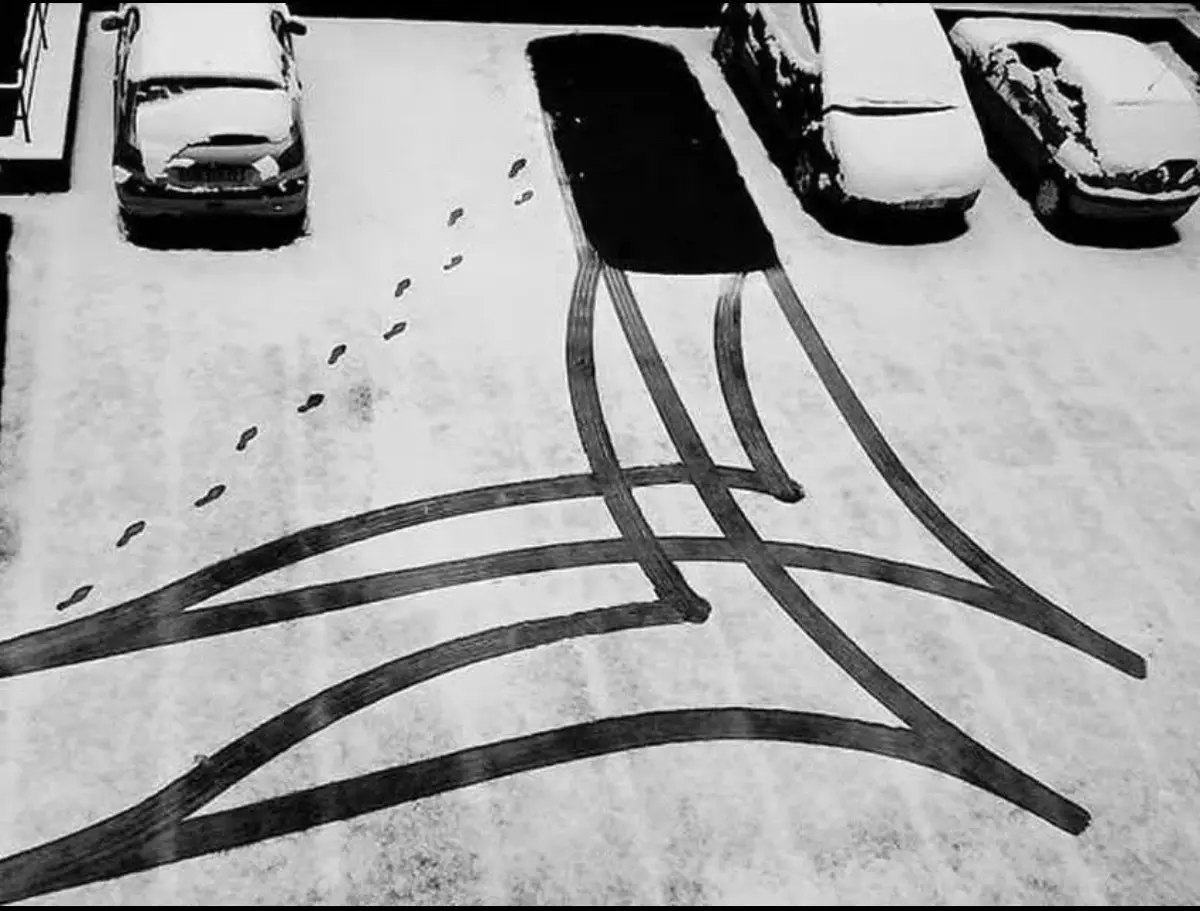 An overhead black-and-white photo shows a snowy parking lot with several cars covered in snow. A trail of footprints leads…
