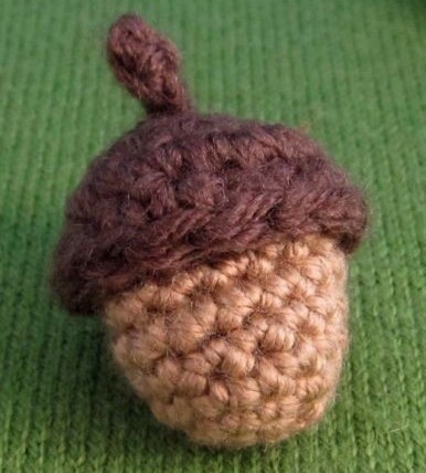 A close-up photo shows a small, hand-crocheted acorn made of brown and tan yarn, resting on a green knitted surface. The…