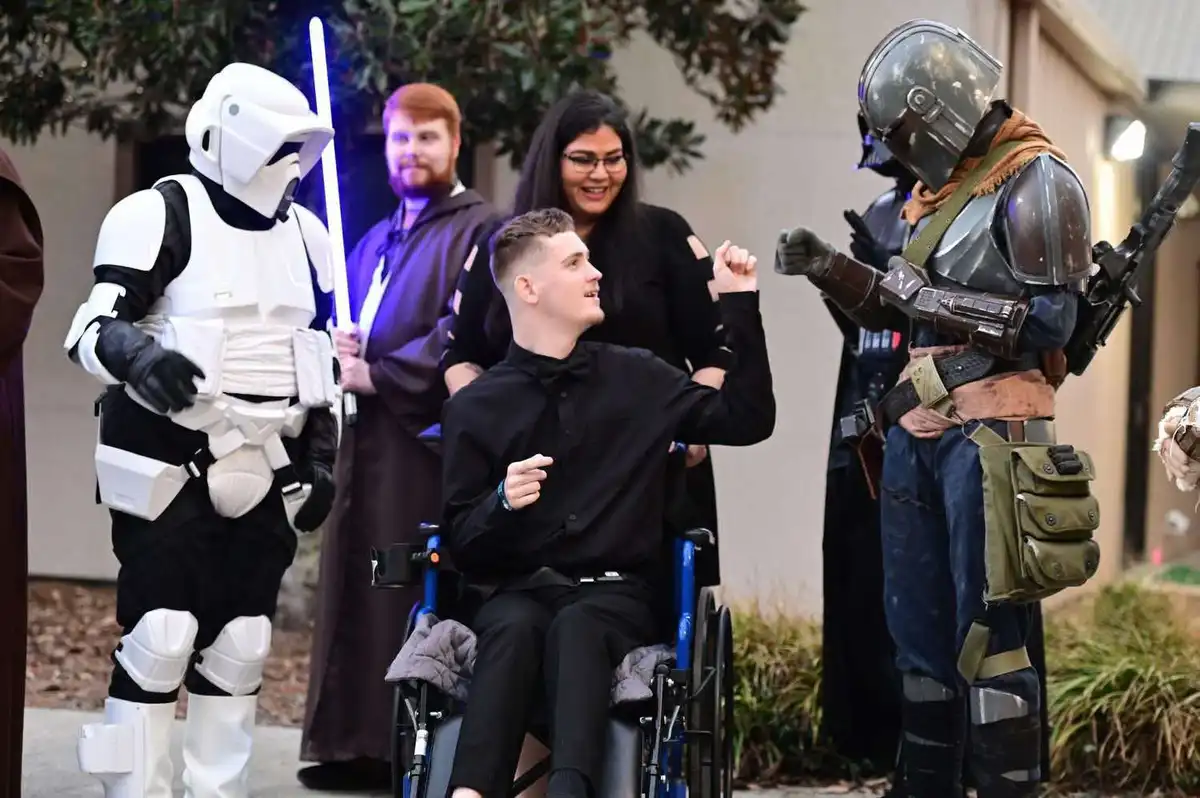 A young man in a wheelchair, dressed in black formal attire, is smiling and raising his fist in celebration while surrounded…