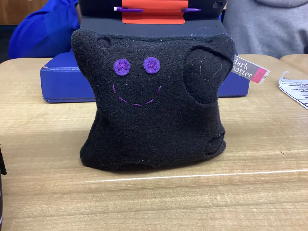 A handmade black felt plush toy with purple embroidered X eyes and a smile sits on a wooden desk, with a tag reading 'dark…
