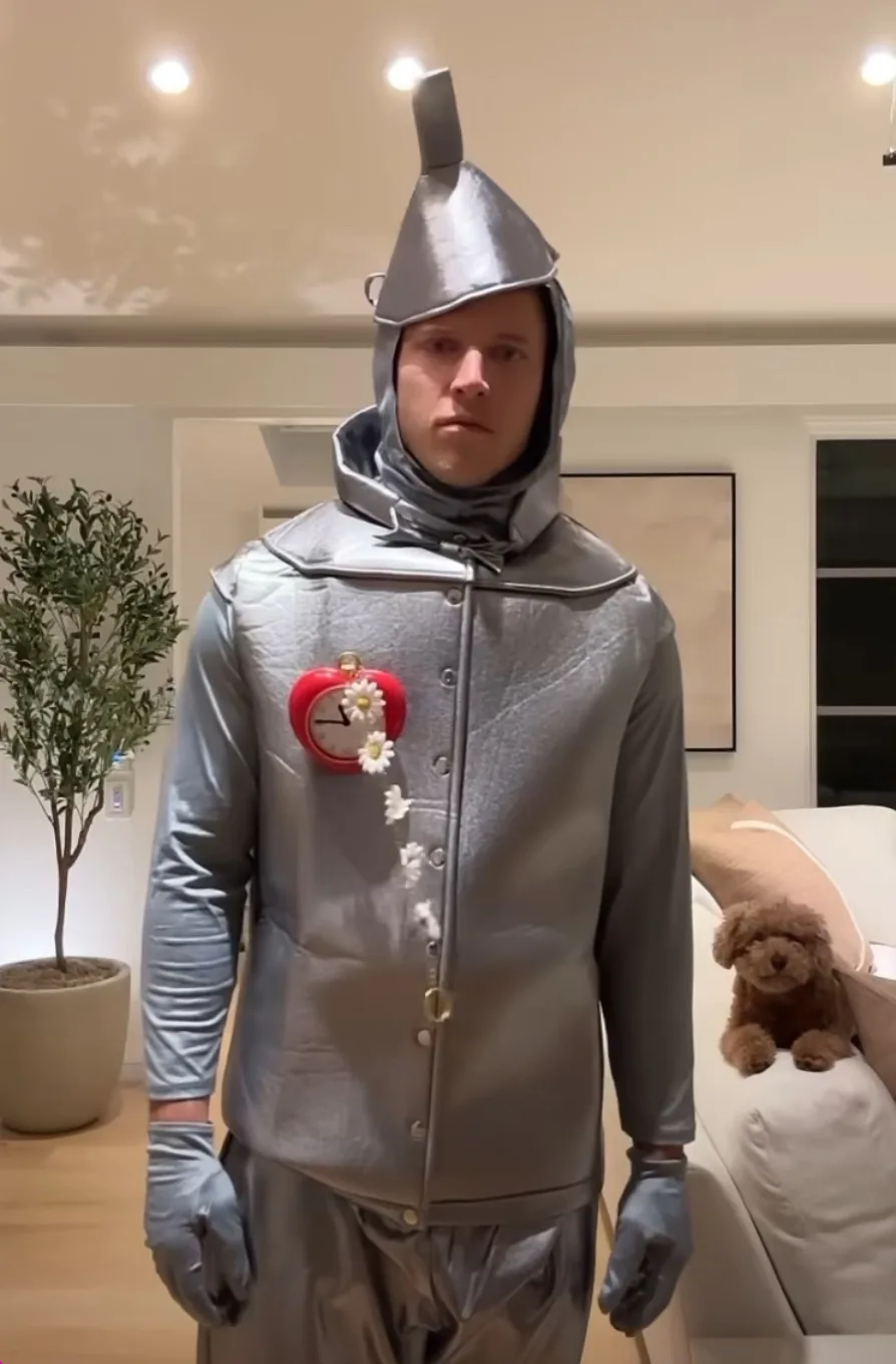 A person dressed in a full silver tin man costume from The Wizard of Oz, complete with a pointed hat, gloves, and a red…