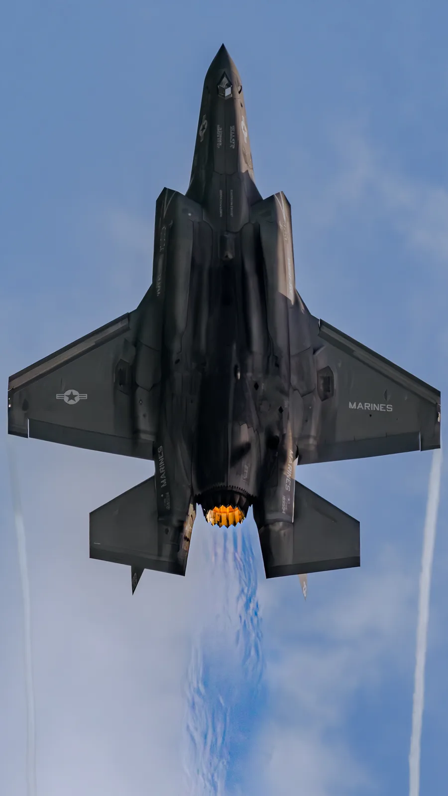 A low-angle shot captures a dark gray F-35 fighter jet flying directly overhead against a blue sky, its afterburner glowing…